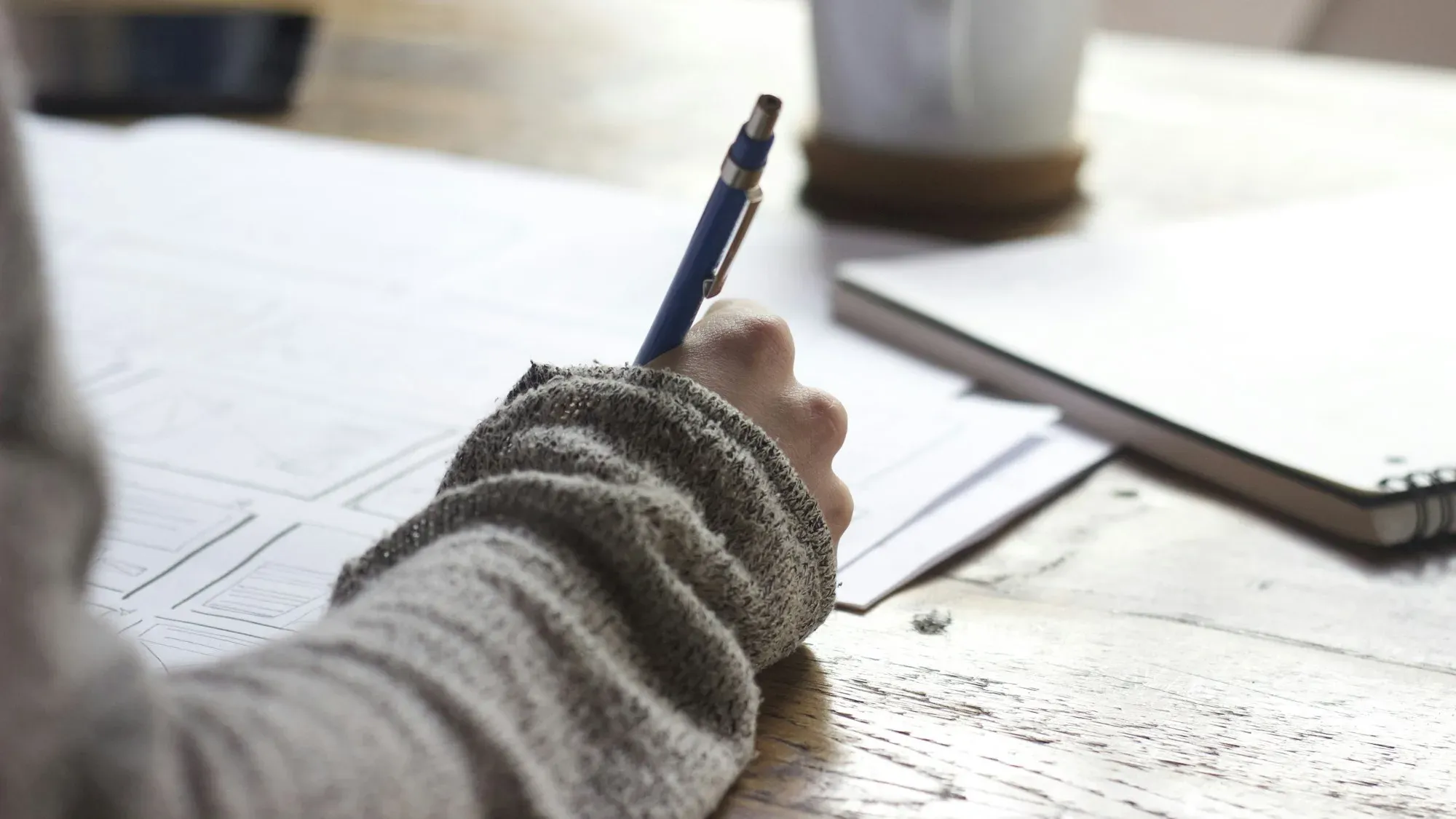 Person taking handwritten notes beside a coffee mug and laptop